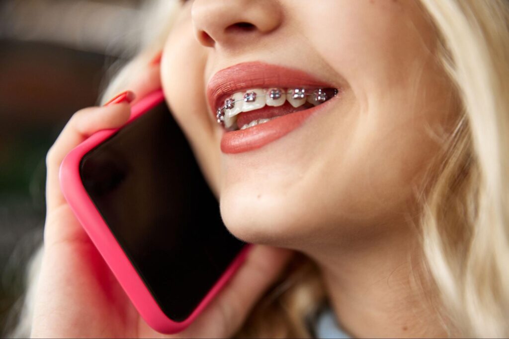 Young woman with braces smiling while talking on a pink smartphone, illustrating the communication aspect of orthodontic treatment at Gross Orthodontics.