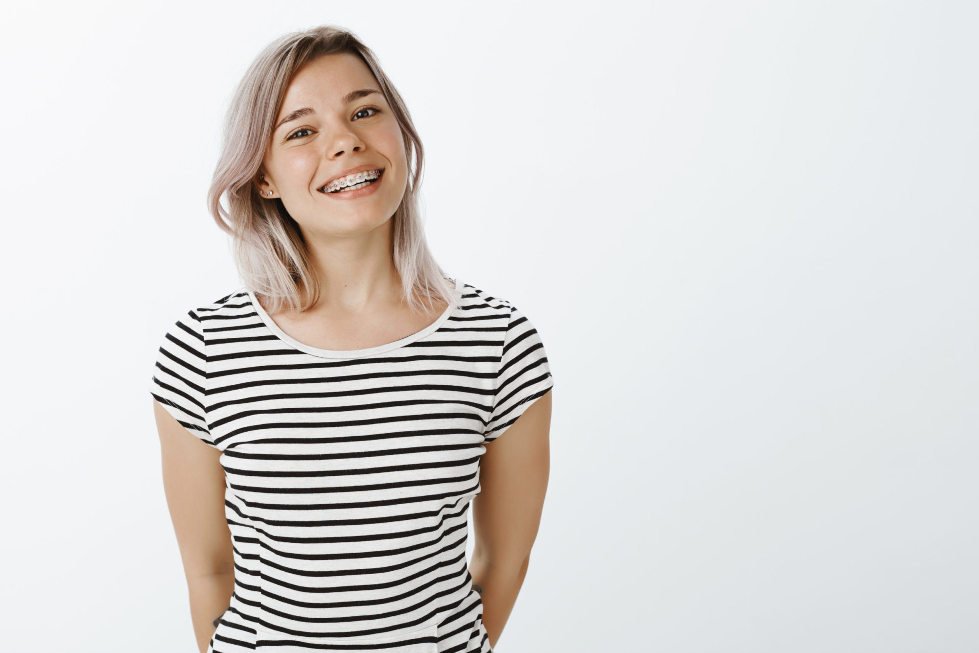 Studio shot of cute caucasian girlfriend with blond hair and braces on teeth standing in casual striped t-shirt and smiling broadly at camera, holding hands behind, enjoying great sunny day Orthodontics El Paso